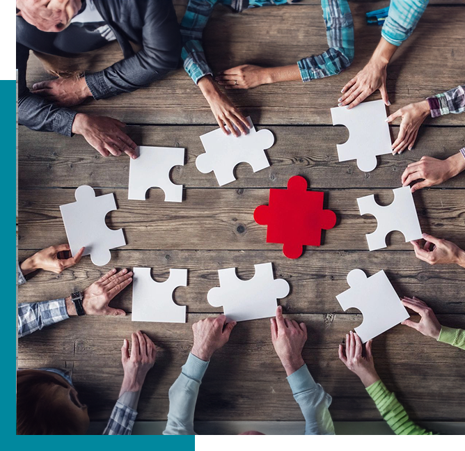 A group of people sits around a wooden table, working together to assemble a puzzle with one red central piece and several white pieces.