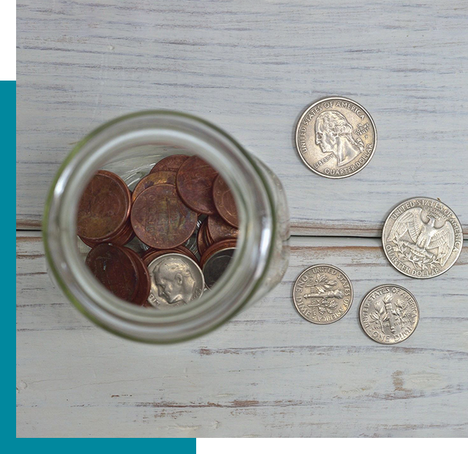 A glass jar filled with pennies stands on a wooden surface. Beside the jar are four quarters and two nickels, spread on the same surface.