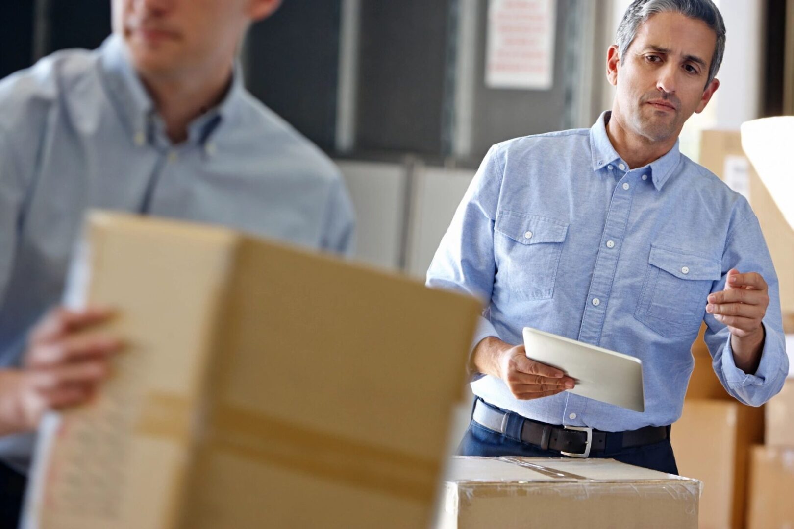 Two men sitting at a table with boxes in the background.